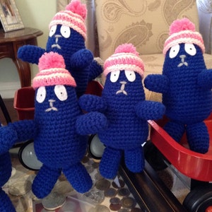May include: A group of handmade blue crochet figures with white eyes, small mouths, and pink and white striped hats. Some figures are in a red wagon. The figures have arms and legs and are standing on a glass surface.
