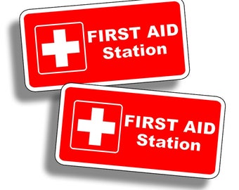 Pode incluir: Dois sinais retangulares vermelhos com bordas brancas. Cada sinal tem um símbolo de cruz branca e o texto "FIRST AID STATION" em branco.