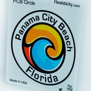 Könnte beinhalten: Weißer Aufkleber mit einem schwarzen Kreis, der ein blaues und orangefarbenes Wellendesign enthält. Der Text "Panama City Beach Florida" ist in einem schwarzen Kreis um die Welle geschrieben. Der Text "Realsticky.com" befindet sich in der oberen rechten Ecke. Der Text "Original PCB Circle" befindet sich in der oberen linken Ecke. Der Text "Made in USA" befindet sich in der unteren linken Ecke. Der Text "Peel Here" befindet sich in der unteren rechten Ecke mit einem Pfeil, der darauf zeigt.