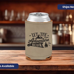 May include: Beige can cooler with a black graphic that says "Let the Adventure Begin" with a mountain and pine tree design.