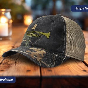 May include: A camouflage baseball cap with a mesh back and a yellow trumpet embroidered on the front. The cap has a curved brim and the text "Ships Next Day" is visible. The text "5 Colors Available" is also visible.