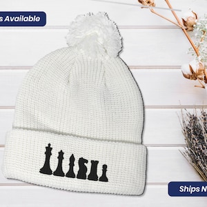 May include: White knit beanie with a white pom pom on top and a black chess piece design on the cuff.  The beanie is on a white wood background.