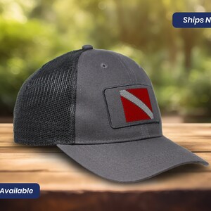 May include: Gray and black mesh trucker hat with a red and white scuba diving flag patch. The hat has a curved brim and a mesh back.