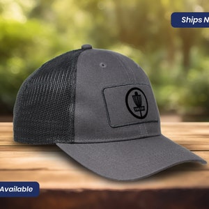 May include: A gray trucker hat with a black mesh back and a curved brim. The hat features a rectangular patch with a black disc golf basket logo. The text "2 Colors Available" is visible.