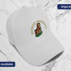 May include: White baseball cap with an embroidered squirrel design and the text "Squirrel Whisperer" in gold lettering.