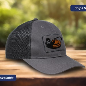 May include: A gray and black mesh trucker hat with a patch of a person shooting a clay pigeon. The patch is embroidered with orange and brown thread. The hat has a curved brim.