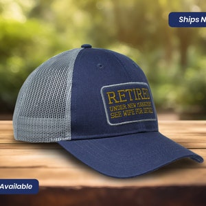 May include: A navy blue and gray mesh trucker hat with a gold embroidered patch that reads "Retired Under New Management See Wife For Details".