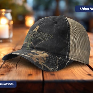 May include: A black and green camouflage trucker hat with a brown mesh back. The hat has the text "Nobody Knows I'm a Lesbian" embroidered on the front.