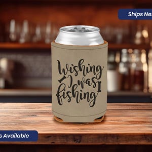 May include: A tan can cooler with the text "Wishing I was fishing" in black lettering. The cooler has a black fishing hook symbol on either side of the text.