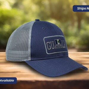 May include: A blue and gray trucker hat with a white mesh back. The hat has a patch on the front that says "GUATEMALA" with the Guatemalan flag.