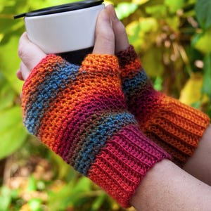May include: A pair of colorful fingerless gloves with a striped pattern in shades of orange, red, brown, and blue. The gloves are crocheted and are being worn by a person holding a white mug.