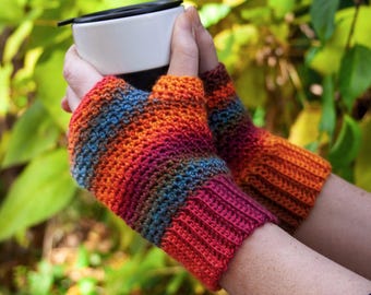 May include: A pair of colourful fingerless gloves with a striped pattern in shades of orange, red, brown, and blue. The gloves are crocheted and are being worn by a person holding a white mug.