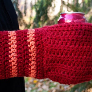 Puede incluir: Un calientamanos de crochet rojo con una franja de color melocotón. El calientamanos está sosteniendo una lata de aluminio roja.