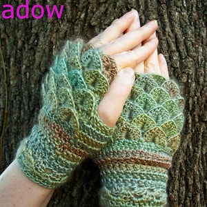 May include: A pair of fingerless crochet gloves featuring a textured scale pattern in variegated shades of green, brown, and tan. The gloves have a ribbed cuff and a scalloped design on the back of the hand. The word Meadow is printed in pink.