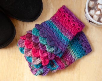 May include: A pair of fingerless gloves crocheted with a variegated yarn in shades of magenta, purple, and teal. The gloves feature a textured scale-like pattern on the exterior, created with overlapping crochet stitches, and a ribbed cuff.