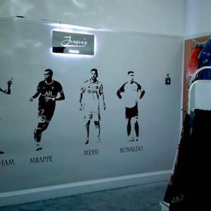 May include: A wall decorated with black silhouette decals of soccer players, including Mbappe, Messi, and Ronaldo. The word "Jenson's" is illuminated on a rectangular sign above the decals. The wall is painted a light gray color.