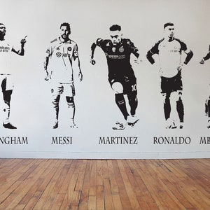 May include: Black and white wall decals of five soccer players: Bellingham, Messi, Martinez, Ronaldo, and Mbappe. Each player is depicted in a dynamic pose, with their names printed below. The decals are on a white wall above a wooden floor.
