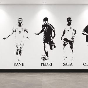 Puede incluir: Un vinilo decorativo en blanco y negro que muestra a cinco jugadores de fútbol en poses de acción. Los nombres Saliba, Kane, Pedri, Saka y Odegaard están impresos en una fuente serif debajo de cada figura individual en la pared.