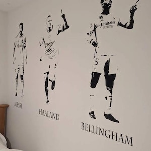May include: Black and white wall decals of soccer players Messi, Haaland, and Bellingham. The players are depicted in action poses, with their names printed below each figure. The Emirates Fly Better logo is visible on Bellingham's jersey.