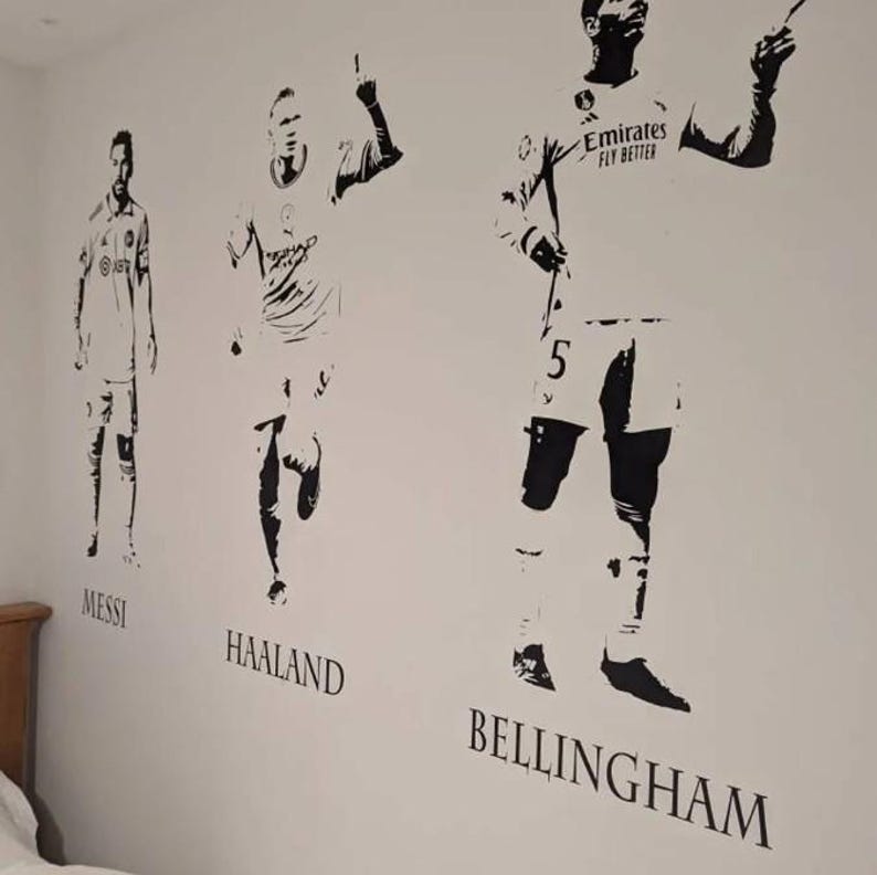 May include: Black and white wall decals of soccer players Messi, Haaland, and Bellingham. The players are depicted in action poses, with their names printed below each figure. The Emirates Fly Better logo is visible on Bellingham's jersey.