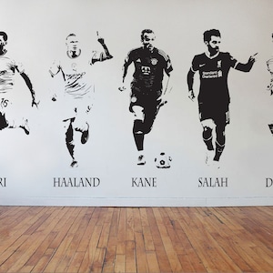 May include: Black and white wall decals featuring five soccer players in action poses. Each player is labeled with their name: Rodri, Haaland, Kane, Salah, and De Bruyne. The decals are applied to a white wall above a wooden floor.