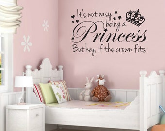 May include: A pink wall with a black vinyl decal that reads "It's not easy being a princess, but hey, if the crown fits". The decal features a crown and hearts. A white bed with a white nightstand and a lamp is in the foreground.