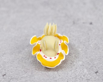 Puede incluir: Una pequeña figura esculpida de un nudibranquio. La criatura presenta un cuerpo de color crema pálido con dos rinóforos erguidos, un borde del manto amarillo brillante ondulado y una serie de pequeños puntos rojos a lo largo del borde blanco.