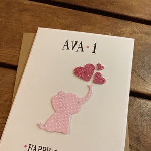 Puede incluir: Un elefante rosa con lunares blancos que sostiene corazones de purpurina rosa con el texto "AVA - 1" en una tarjeta blanca.