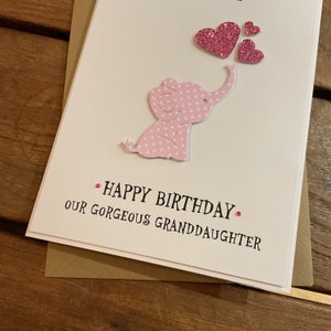Puede incluir: Un elefante rosa con lunares blancos y un coraz&oacute;n rosa sobre su cabeza. El texto "Happy Birthday Our Gorgeous Granddaughter" est&aacute; escrito debajo del elefante.