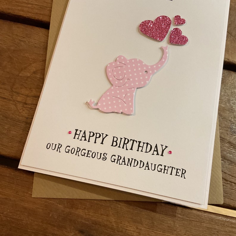 Puede incluir: Un elefante rosa con lunares blancos y un coraz&oacute;n rosa sobre su cabeza. El texto "Happy Birthday Our Gorgeous Granddaughter" est&aacute; escrito debajo del elefante.