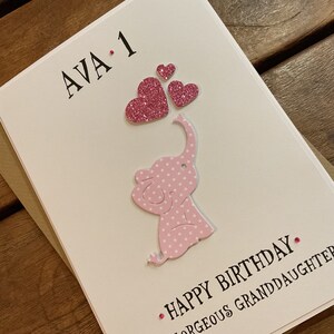 Puede incluir: Una tarjeta blanca con un elefante rosa con lunares blancos que sostiene corazones rosas con purpurina con el texto "AVA. 1" y "FELIZ CUMPLEA&Ntilde;OS".