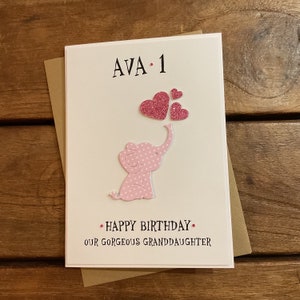 Puede incluir: Una tarjeta de cumplea&ntilde;os rosa con un elefante rosa sosteniendo corazones de purpurina rosa. La tarjeta dice "AVA. 1" y "Feliz cumplea&ntilde;os. Nuestra hermosa nieta".
