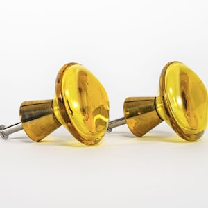 May include: Two cabinet knobs featuring round, translucent yellow glass heads attached to conical, brushed brass-toned metal bases. Each knob includes a short, silver-toned metal mounting screw extending from the back of the base.