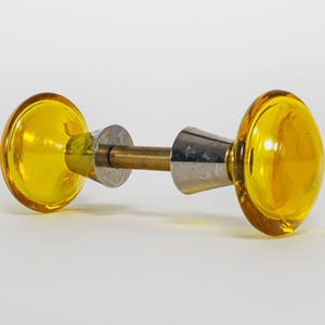 Puede incluir: Un par de pomos de puerta vintage con un diseño de vidrio redondeado de color amarillo brillante. Los pomos están conectados por una varilla de latón y herrajes de metal plateado. El vidrio tiene un acabado brillante.