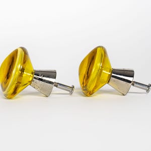 May include: Two yellow glass cabinet knobs with a silver-colored metal base and screw. The knobs have a rounded, asymmetrical shape and a swirled pattern within the yellow glass. The hardware is set against a white background.