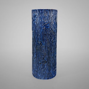 Könnte beinhalten: Eine hohe, zylindrische Vase in tiefblau. Die Vase hat eine strukturierte Oberfläche mit vertikalen Linien und geprägten Fischmotiven. Das Design verleiht der Vase ein einzigartiges, handwerkliches Aussehen, geeignet für die Inneneinrichtung.