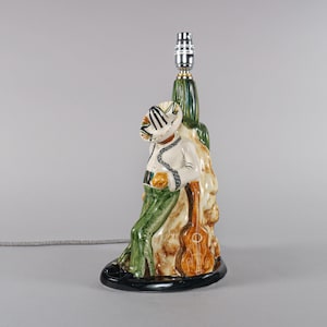 Könnte beinhalten: Eine Keramiklampe in Form eines Mariachi-Musikers. Die Lampe zeigt eine Figur in einem Sombrero, einem weißen Hemd, grünen Hosen und einer Gitarre. Der Sockel ist schwarz, und die Lampe hat ein grünes und braunes Farbschema.