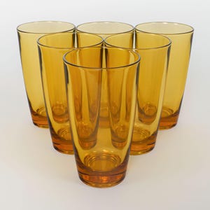 Op de afbeelding: Zes amberkleurige glazen tumblers, elk met een afgeronde bodem en rechte zijden, gerangschikt in een cirkel op een wit oppervlak.