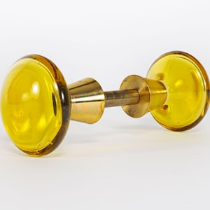 May include: A door knob set featuring two round, translucent yellow glass handles connected by a central metallic brass spindle and conical base plates. The design consists of smooth, polished surfaces with a clear, vibrant yellow color profile.