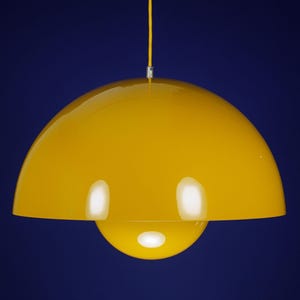 Op de afbeelding: Een heldergele hanglamp met een afgeronde, koepelvormige kap en een kleiner, bolvormig onderste gedeelte. Hij hangt aan een gele kabel tegen een diepblauwe achtergrond. Het ontwerp is modern en minimalistisch.