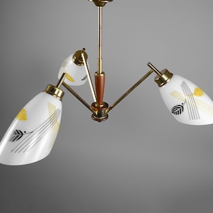 Könnte beinhalten: Eine Vintage-Deckenleuchte mit drei weißen Glasschirmen. Jeder Schirm ist mit gelben und schwarzen Blatt- und Liniendesigns verziert. Die Leuchte hat Messing- und Holzakzente und ist an einer Messingstange aufgehängt.