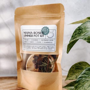 May include: A brown paper bag with a clear window showing the contents of a Mama Roses Simmer Pot Kit. The kit includes orange, cinnamon, cardamom pods, cedar, salt, clove, and cranberry. The bag has a label with the text "Mama Roses Simmer Pot Kit" and "winter hot".