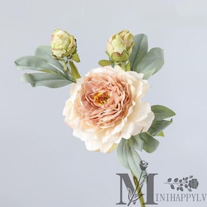 May include: A single artificial peony flower with a large, layered peach-colored bloom and a yellow center. Two green buds and several gray-green leaves are also visible. The flower is set against a light gray background.