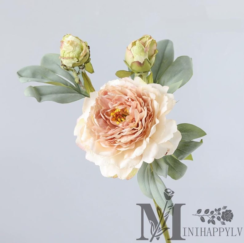 May include: A single artificial peony flower with a large, layered peach-colored bloom and a yellow center. Two green buds and several gray-green leaves are also visible. The flower is set against a light gray background.