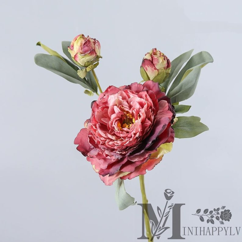 May include: A single artificial peony flower with a large, multi-layered, deep pink bloom. Two smaller buds and several green leaves are also visible. The stem is a light green color. The flower is set against a plain white background.