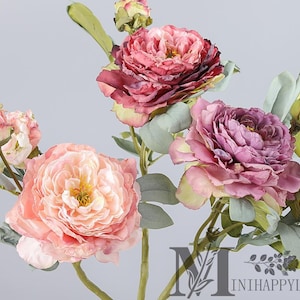 May include: Close-up of artificial peony flowers in shades of pink, peach, and purple. The flowers have multiple layers of petals and are surrounded by green leaves and stems. The overall aesthetic is soft and romantic.