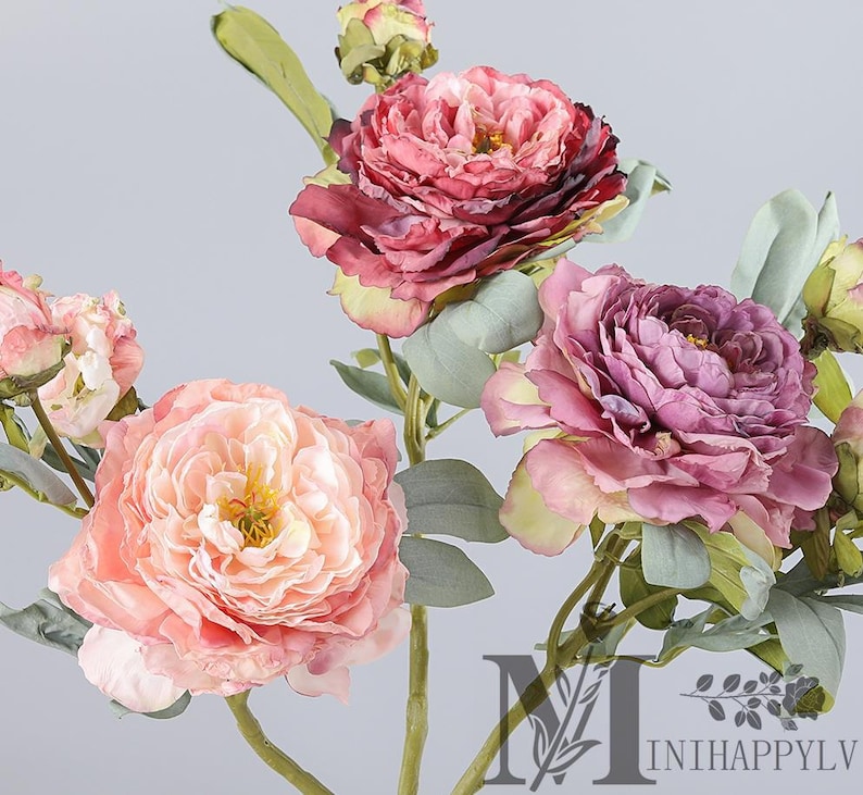 May include: Close-up of artificial peony flowers in shades of pink, peach, and purple. The flowers have multiple layers of petals and are surrounded by green leaves and stems. The overall aesthetic is soft and romantic.