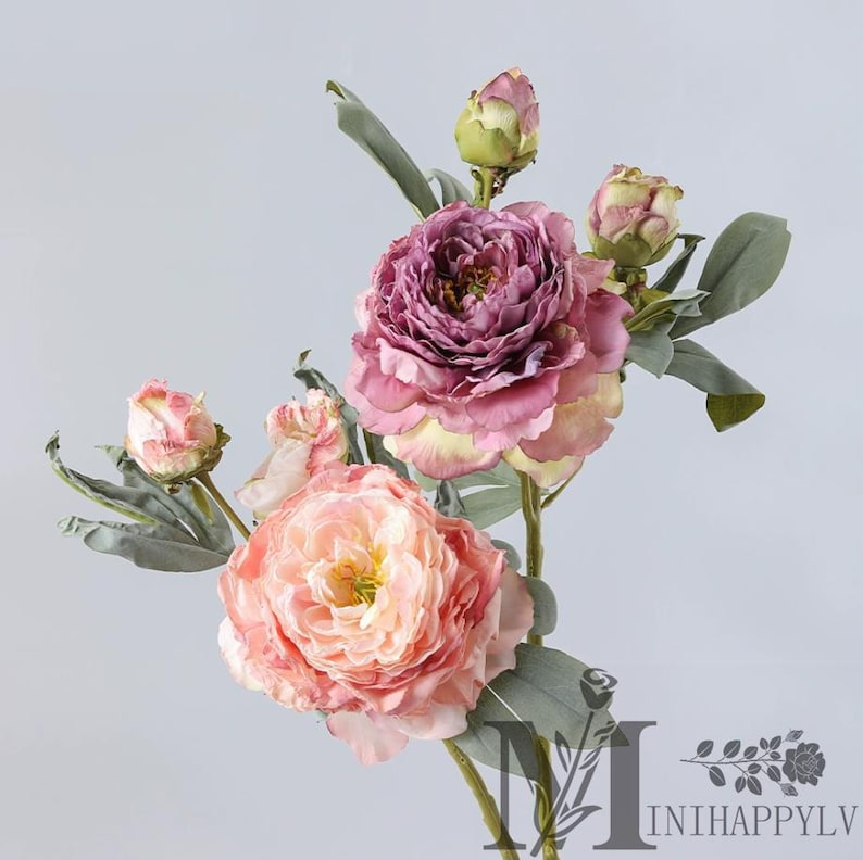 May include: A bouquet of artificial peonies in shades of pink, peach, and purple. The flowers have multiple layers of petals and are accompanied by green leaves and buds. The arrangement is set against a light gray background.