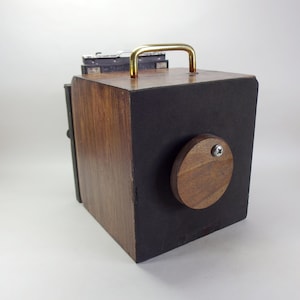 May include: A box-shaped camera constructed from dark wood and black panels. It features a gold-toned metal handle on the top, a circular wooden lens cover secured with a silver screw on the front, and a black metal film back mechanism at the rear.