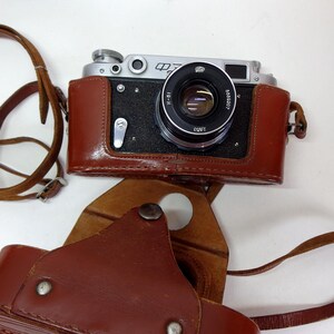 Puede incluir: Una cámara telemétrica vintage de color plateado y negro con un estuche de cuero marrón y una correa a juego. El objetivo presenta texto en cirílico y el cuerpo de la cámara muestra los caracteres cirílicos ФЭД. El estuche es desmontable.